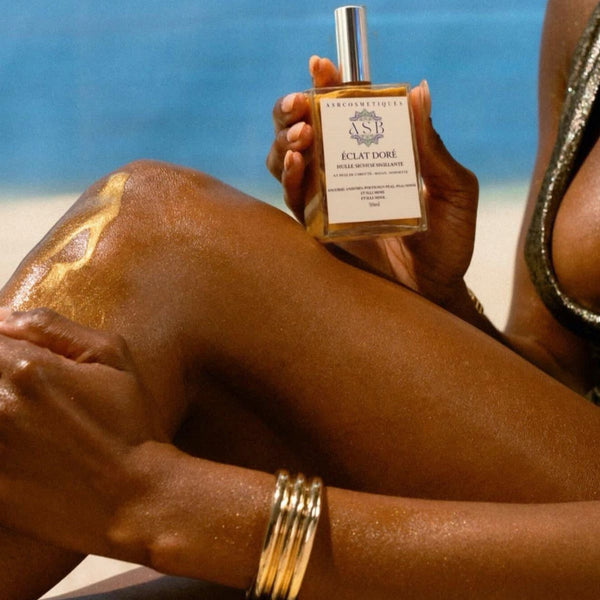 gros plans d'une jambe de femme avec le flacon d'huile scintillante en train de l'appliquer sur sa jambe, dans une main elle tien le flacon et de l'autre elle applique le l'huile